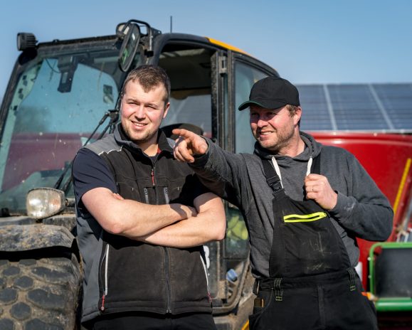 Two men stood infront of a tractor. The sun is shining on their face and they are stood next to eachother. One man has his arms crossed and is looking into the distance, the man next to him is in a cap and overalls and is pointing in the direction the man is looking. They are both smiling subtly.